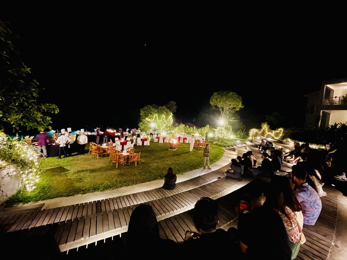 A lawn dinner at night, with guests on amphitheatre steps, a fire pit and chefs at work